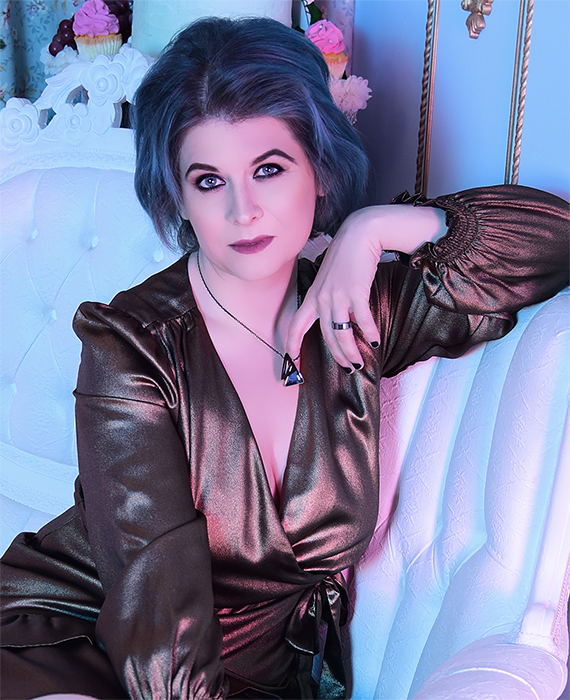 Portrait of Keira North, a nonbinary person with blue hair styled in soft waves, wearing a metallic bronze wrap dress. They are seated on a white, tufted sofa with one arm resting on the back, looking directly at the camera with a confident expression. The setting includes soft, cool lighting that enhances the color of their hair and dress.