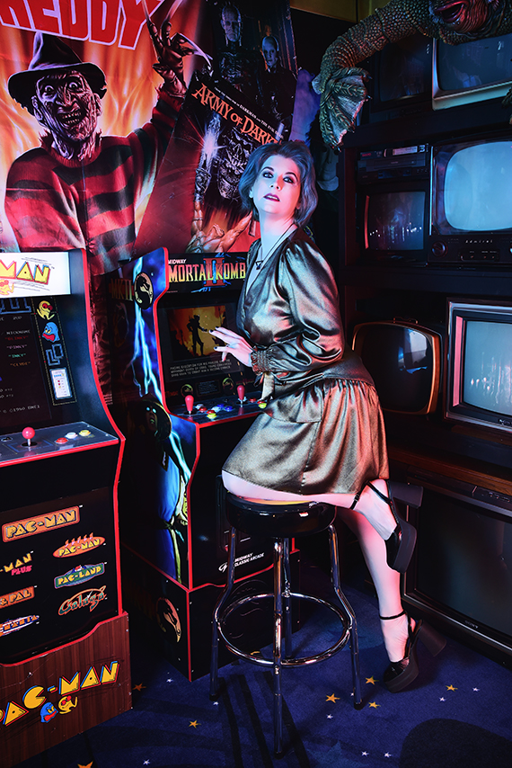 Keira North, a nonbinary individual with blue hair, is seated on a stool in a retro arcade room, playing a Mortal Kombat arcade game. They are wearing a metallic bronze wrap dress and black high heels. The room is decorated with vintage televisions and posters, including one of Freddy Krueger from Nightmare on Elm Street and another for Army of Darkness. The setting is illuminated with neon lights, creating a vibrant, retro-infused atmosphere.